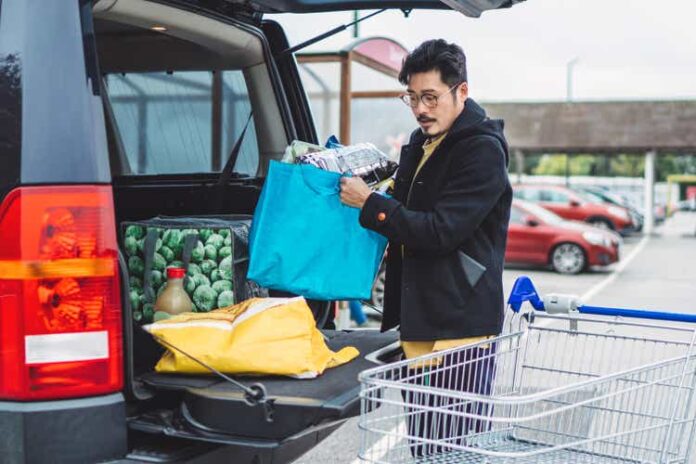 Young Asian man putting away bags of groceries in his car trunk at supermarket car park