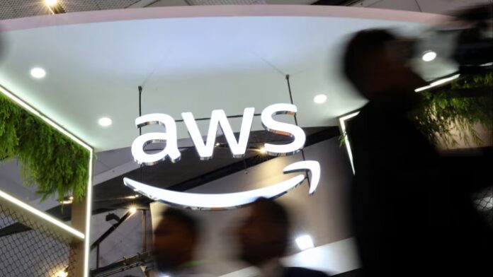 Large illuminated ‘AWS’ logo with Amazon’s smile symbol at an exhibitor stall, with people walking past in the foreground.