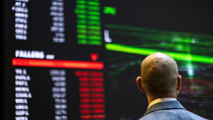 A person in a suit looks at a digital board displaying stock prices, with green and red numbers, at the London Stock Exchange.
