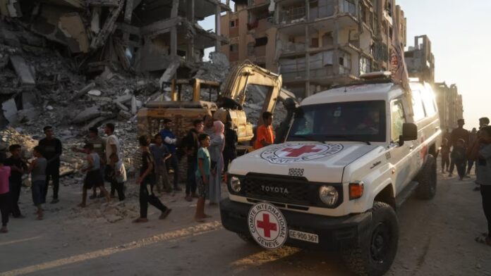 A Red Cross vehicle arrives near a crowd and heavy machinery amid rubble and damaged buildings in Hamad City, Khan Younis.