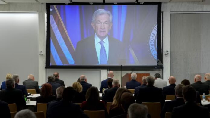 Jerome Powell appears on a large screen, speaking virtually to an audience seated at the Federal Reserve Board Community Bank Conference.