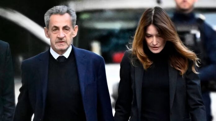 Nicolas Sarkozy and Carla Bruni-Sarkozy walk together, both dressed in dark clothing, with serious expressions.