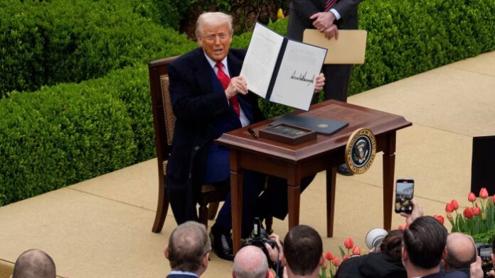 Donald Trump sits at a desk outdoors, holding up a signed executive order during a trade announcement event