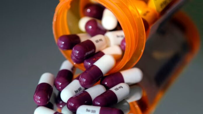 Purple and white capsules labeled &quot;100 mg&quot; spill from an orange prescription bottle onto a reflective surface.