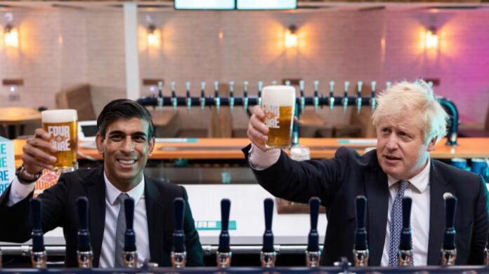 Boris Johnson and Rishi Sunak raise pint glasses of beer behind the bar at Fourpure Brewery, both wearing suits and smiling.