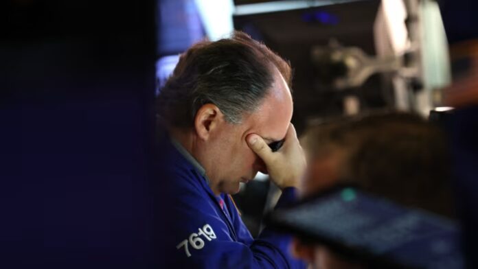 A trader on the New York Stock Exchange floor sits at his desk with his hand covering his face, appearing stressed or deep in thought.