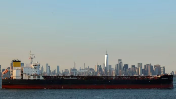 A large cargo ship leaves Port Jersey Container Terminal with the Manhattan skyline visible in the background.