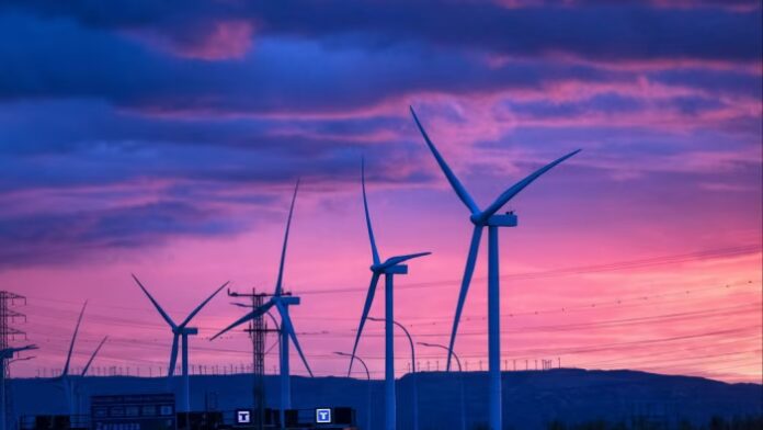 Wind turbines stand against a vibrant purple and pink sunset sky, with power lines and distant hills in Zaragoza, Spain.