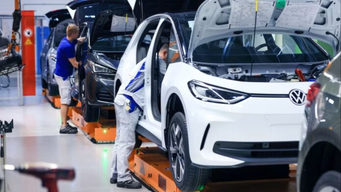Workers assemble Volkswagen ID.3 and Cupra Born electric sedans on a production line, with car hoods open at a Volkswagen plant.