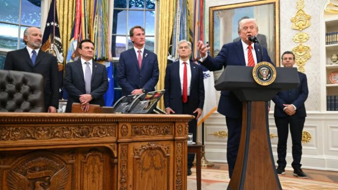 Donald Trump speaks at the Oval Office podium as Howard Lutnick, Pascal Soriot, Glenn Youngkin, Mehmet Oz and Marty Makary stand nearby.