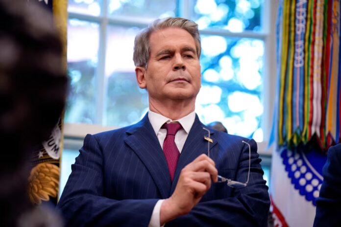 Scott Bessent stands in the Oval Office holding eyeglasses, wearing a pinstripe suit and red tie.