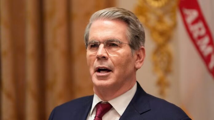 Scott Bessent speaks, mouth open, in a suit and red tie, with a blurred ornate background and part of a banner visible.