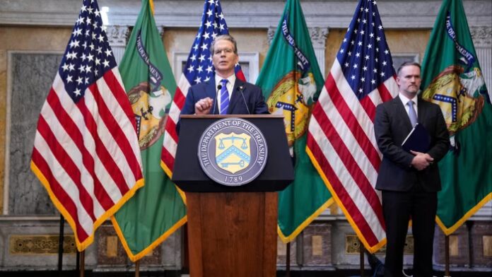 Scott Bessent speaks at a podium with the US Treasury seal as Jamieson Greer stands nearby, both flanked by US and Treasury flags.