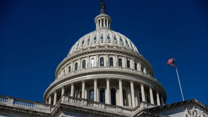 The dome of the US Capitol building against a clear blue sky, with an American flag flying nearby.